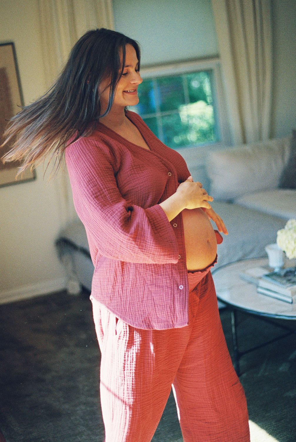 Smiling pregnant woman in pink linen matching set cradling baby bump indoors.