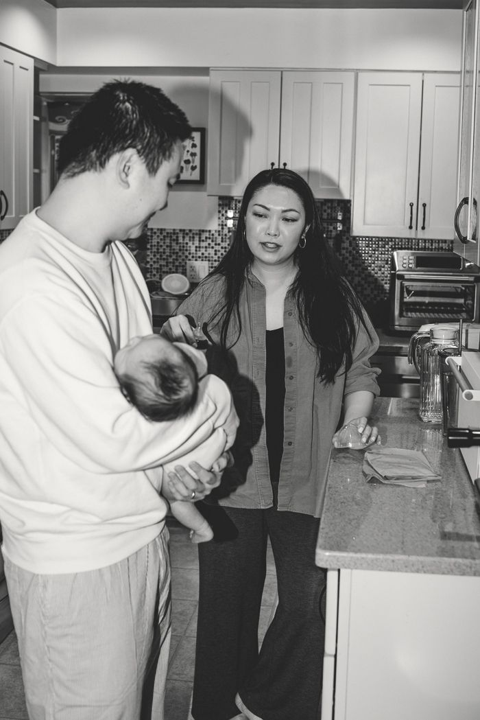 Black and white photo of young Asian couple with newborn baby in modern kitchen home setting.