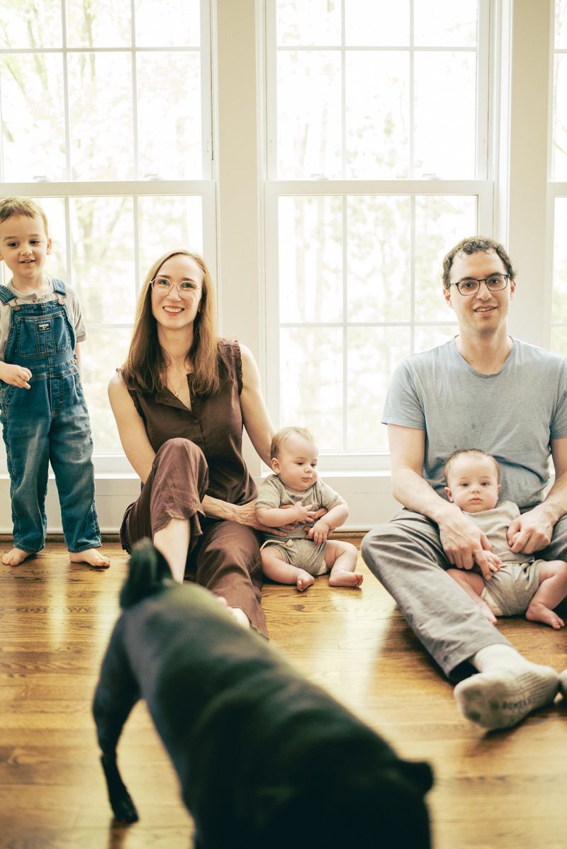 Family of four with two babies and a toddler sitting on hardwood floor near windows, with a black dog in the foreground.