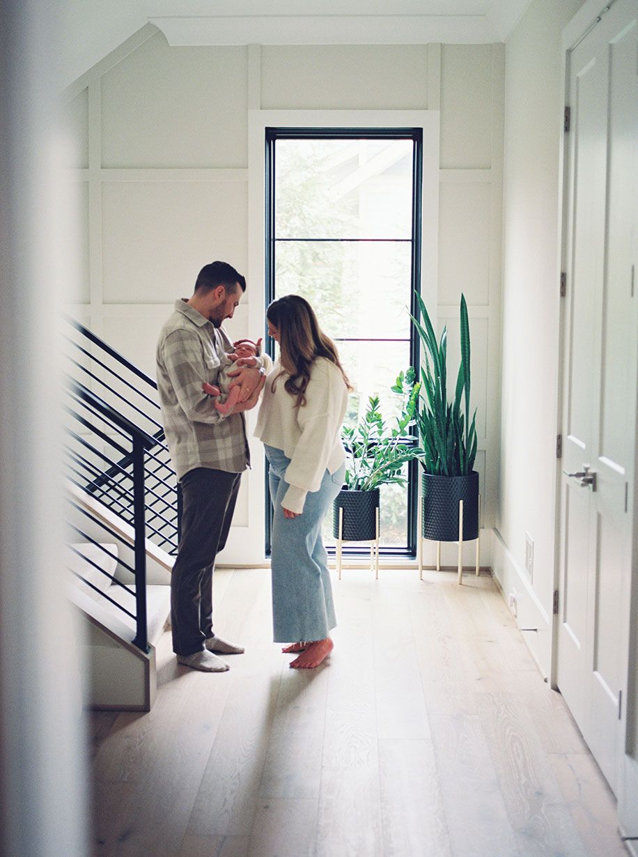 A couple stands in a bright hallway gazing at their newborn baby by a window with indoor plants.