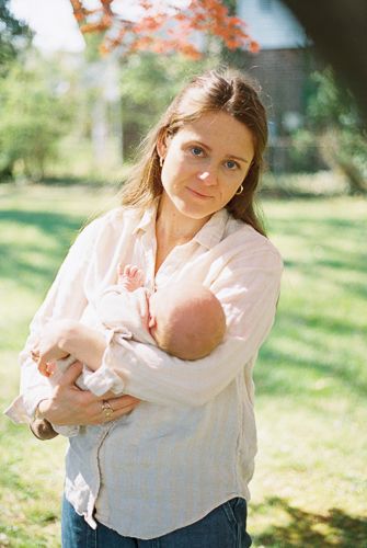 A young mother holds her newborn baby outdoors on a sunny day, standing on green grass with trees in the background.