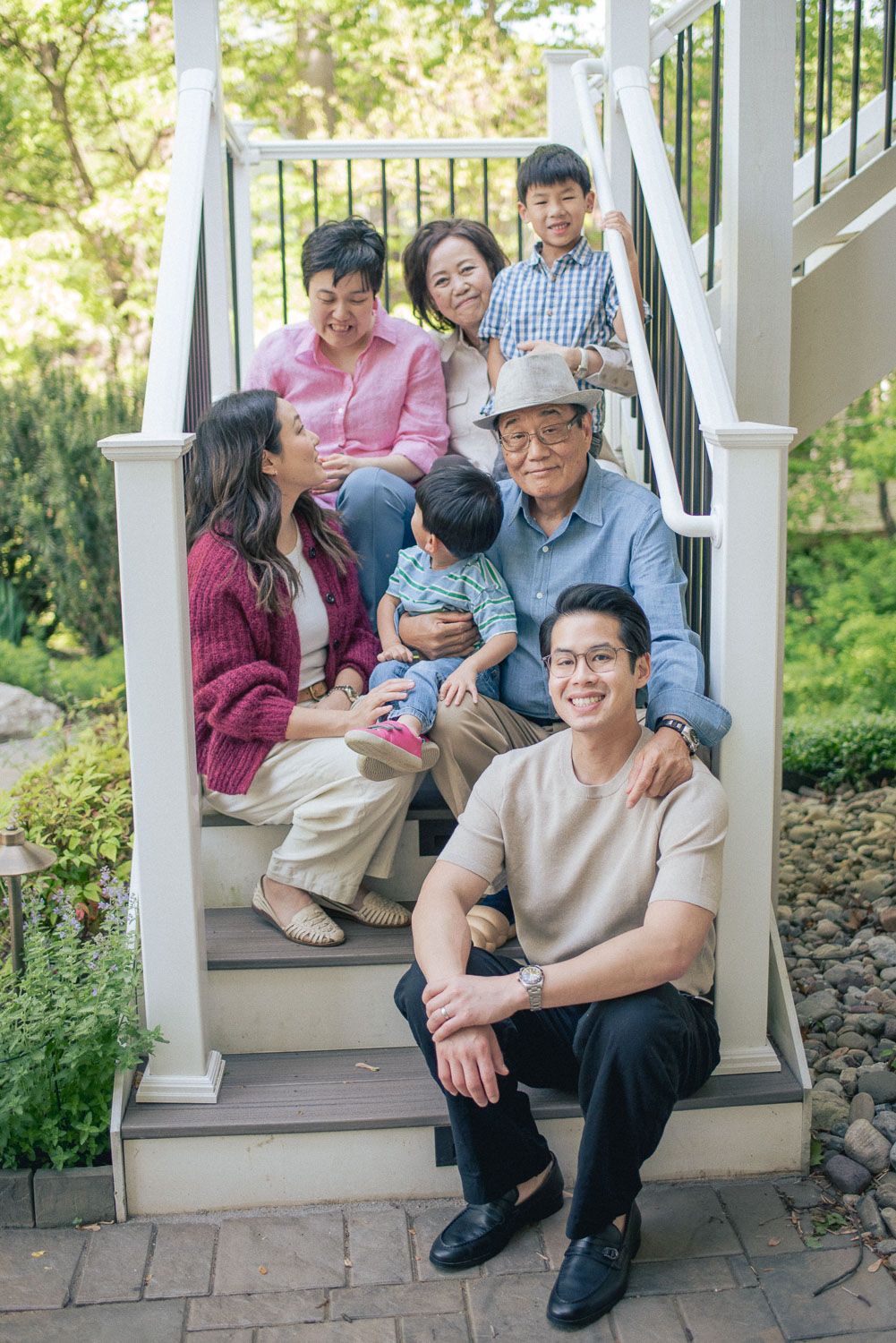 Multi-generational Asian family posing together on outdoor staircase steps surrounded by lush green garden.