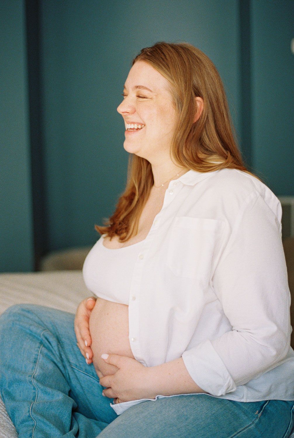 Smiling pregnant woman in white shirt and jeans sitting on bed, cradling baby bump with both hands.