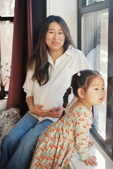 Pregnant Asian woman in white shirt and jeans sits near window with young girl in floral dress with pigtails.