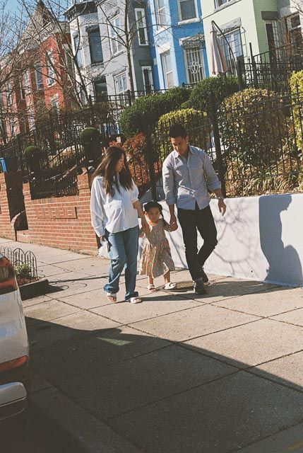Family of three holding hands walking on a sunny urban sidewalk past colorful row houses.