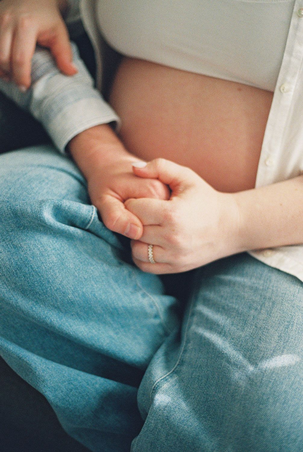Pregnant woman in jeans holding hands with partner, showing baby bump and a delicate ring on her finger.