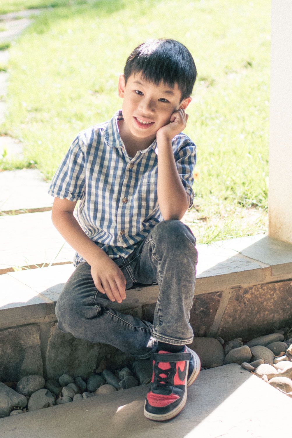 Young Asian boy in plaid shirt and jeans sitting outdoors on stone steps, smiling and resting hand on cheek.