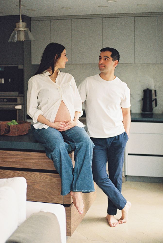 A couple in white shirts and blue jeans sit together on a wooden floor in a minimalist modern interior.