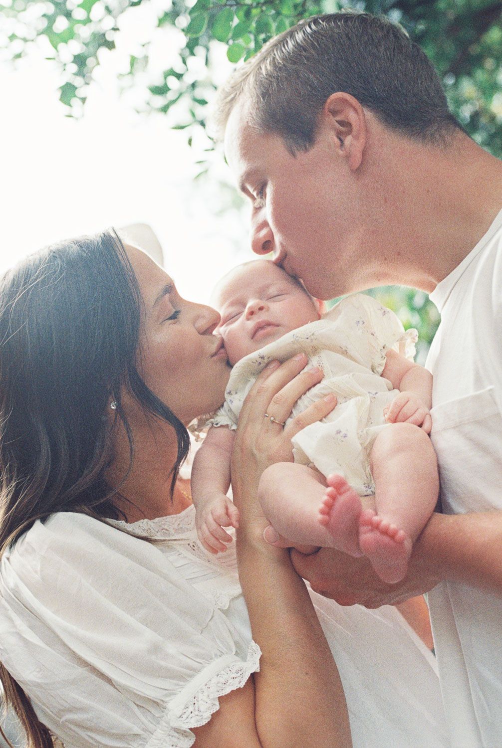 A loving couple in white clothing kisses their newborn baby outdoors under green trees in a tender family portrait.