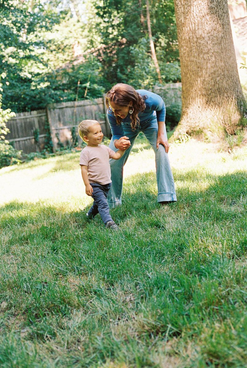 Woman bending down to interact with a toddler boy on a sunny green lawn in a backyard setting.
