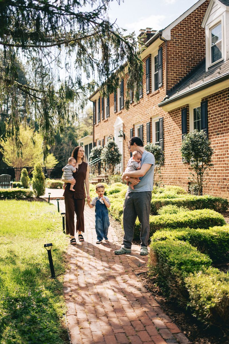 Family of four with two young children standing on brick path outside large brick home on sunny day.