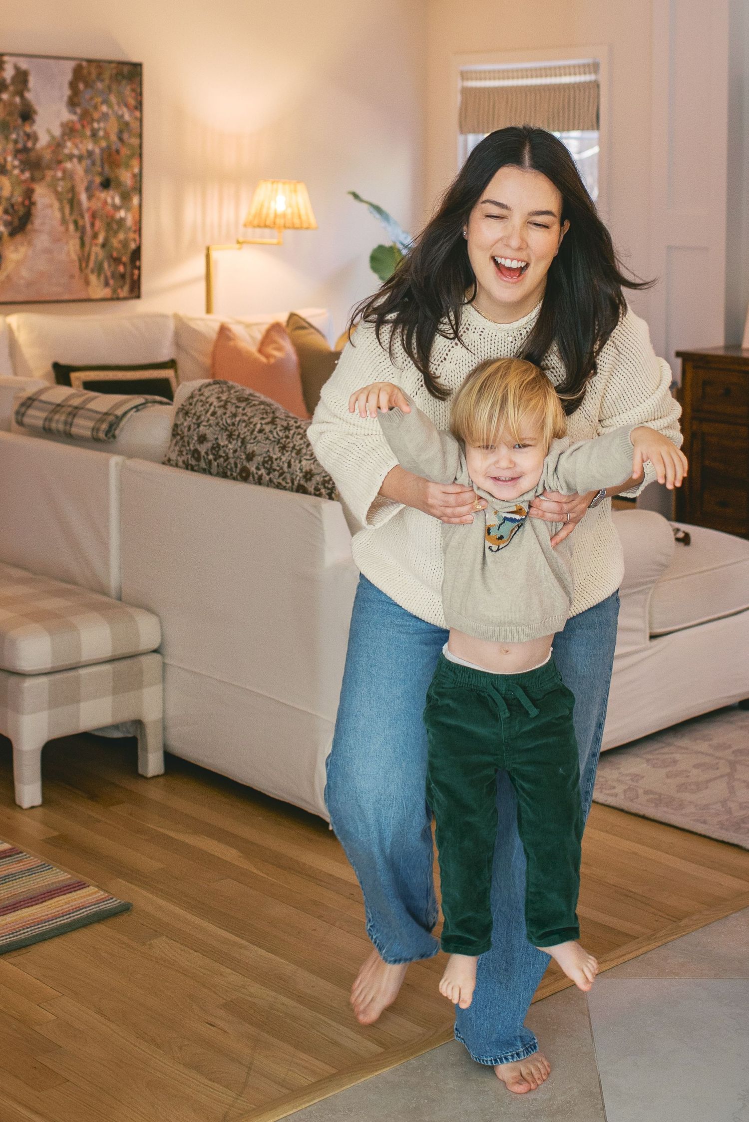 Parent and child playing and laughing together in a bright living room with hardwood floors and white furniture.