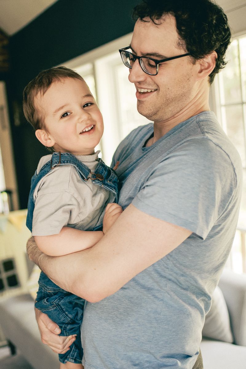 Smiling father in glasses holding laughing toddler boy in denim overalls indoors.