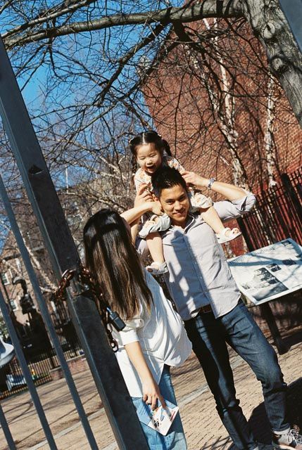 A happy family plays outdoors, with a laughing child sitting on a man's shoulders near a fence.