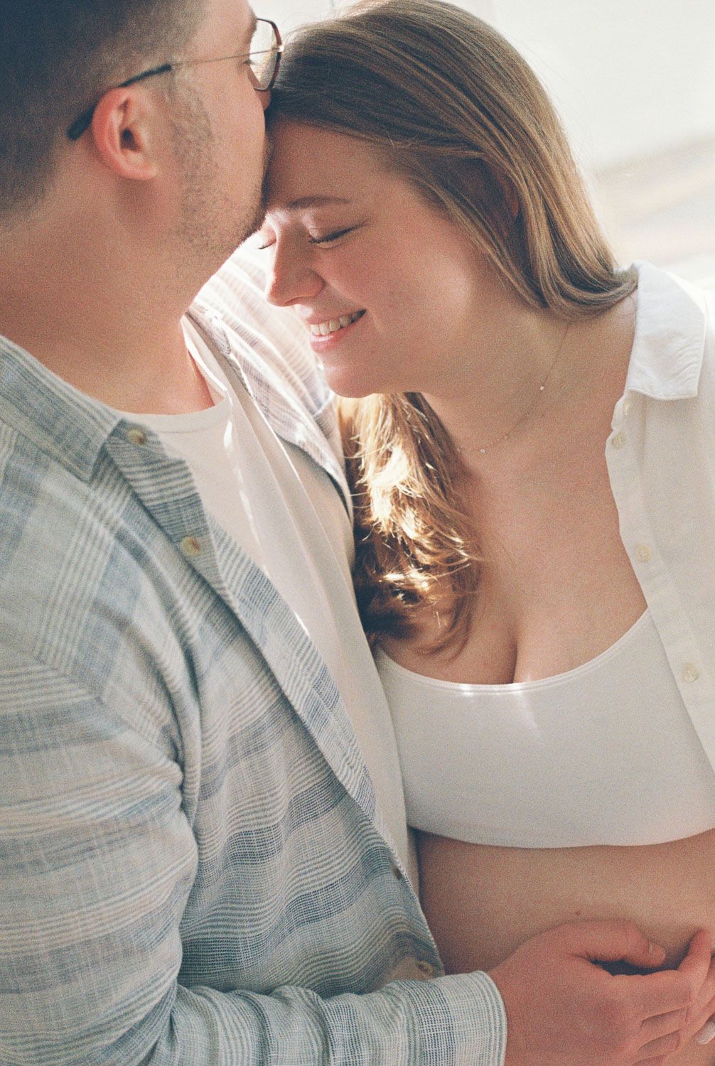 A couple shares an intimate forehead-to-forehead moment, smiling softly during a warm, sunlit maternity photo session.