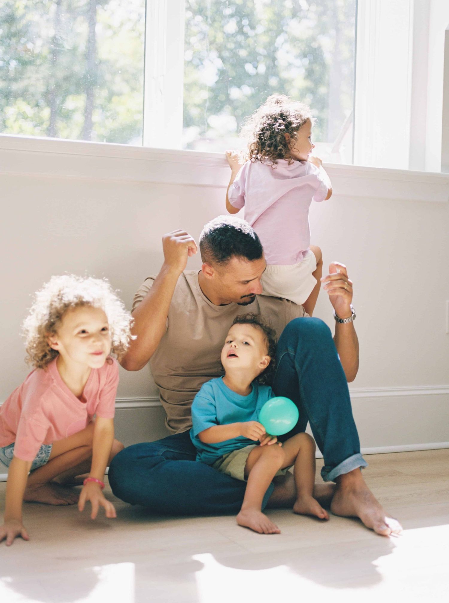 Family members crawl and play together on a sunlit floor near large windows.