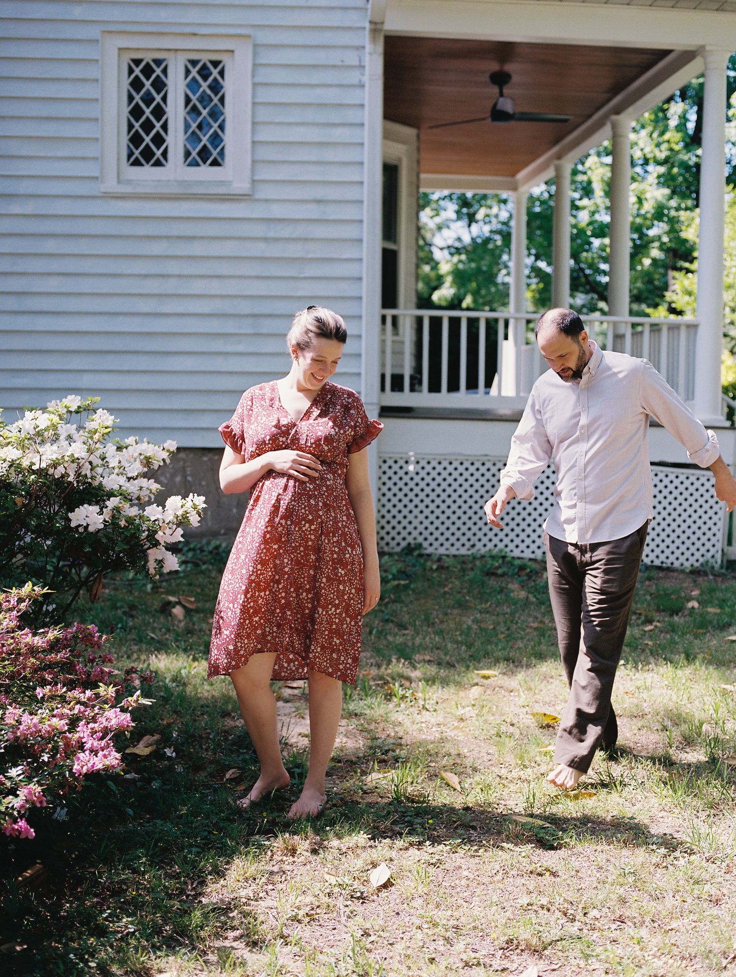 Two people dancing joyfully together on the lawn of a white country house with a wraparound porch in summertime.