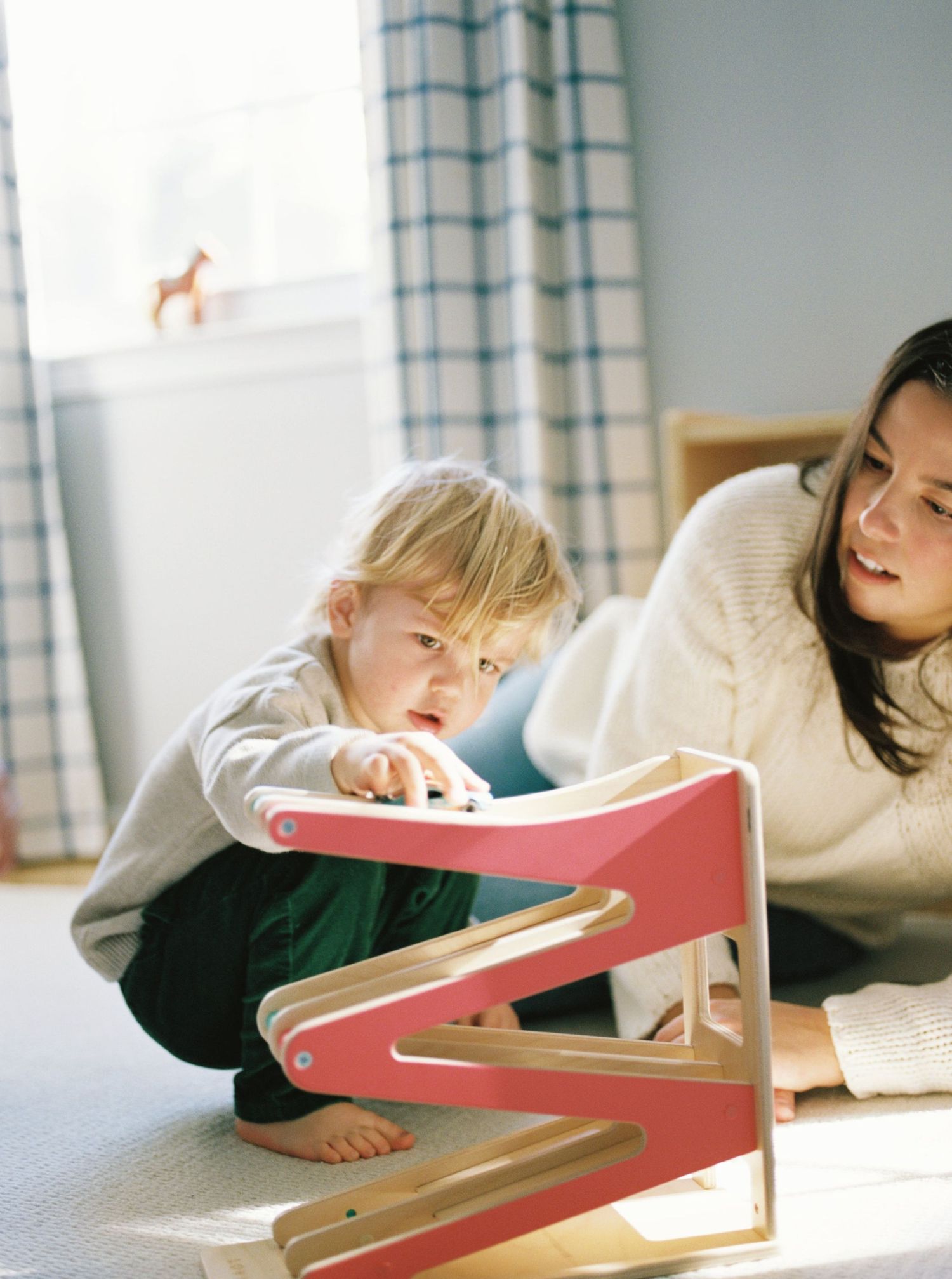 A child lying on the floor plays with a pink wooden letter Z in a room with checkered curtains.