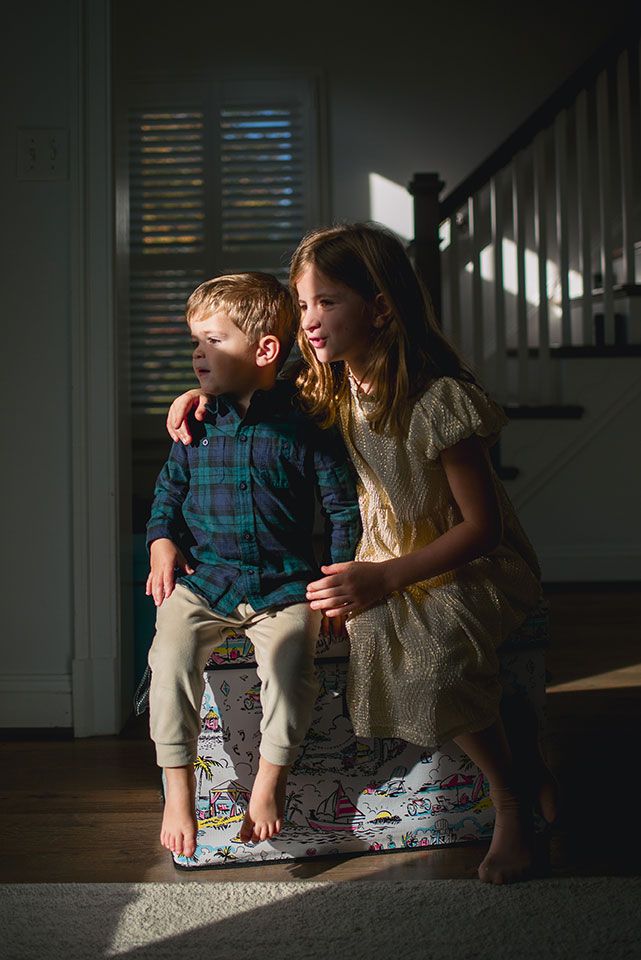 A parent and child sit together in a dimly lit room on a carpet, sharing a tender moment in natural window light.