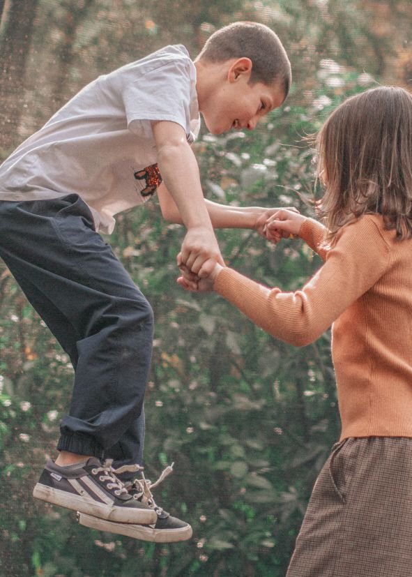 Two people share a joyful moment dancing and jumping together in a garden setting with greenery in the background.