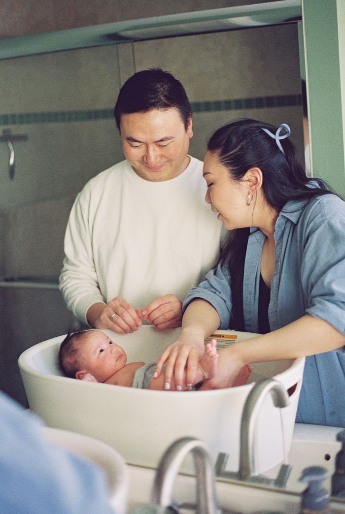 Parents lovingly bathe their newborn baby in a small tub, sharing a tender family moment in a hospital room.