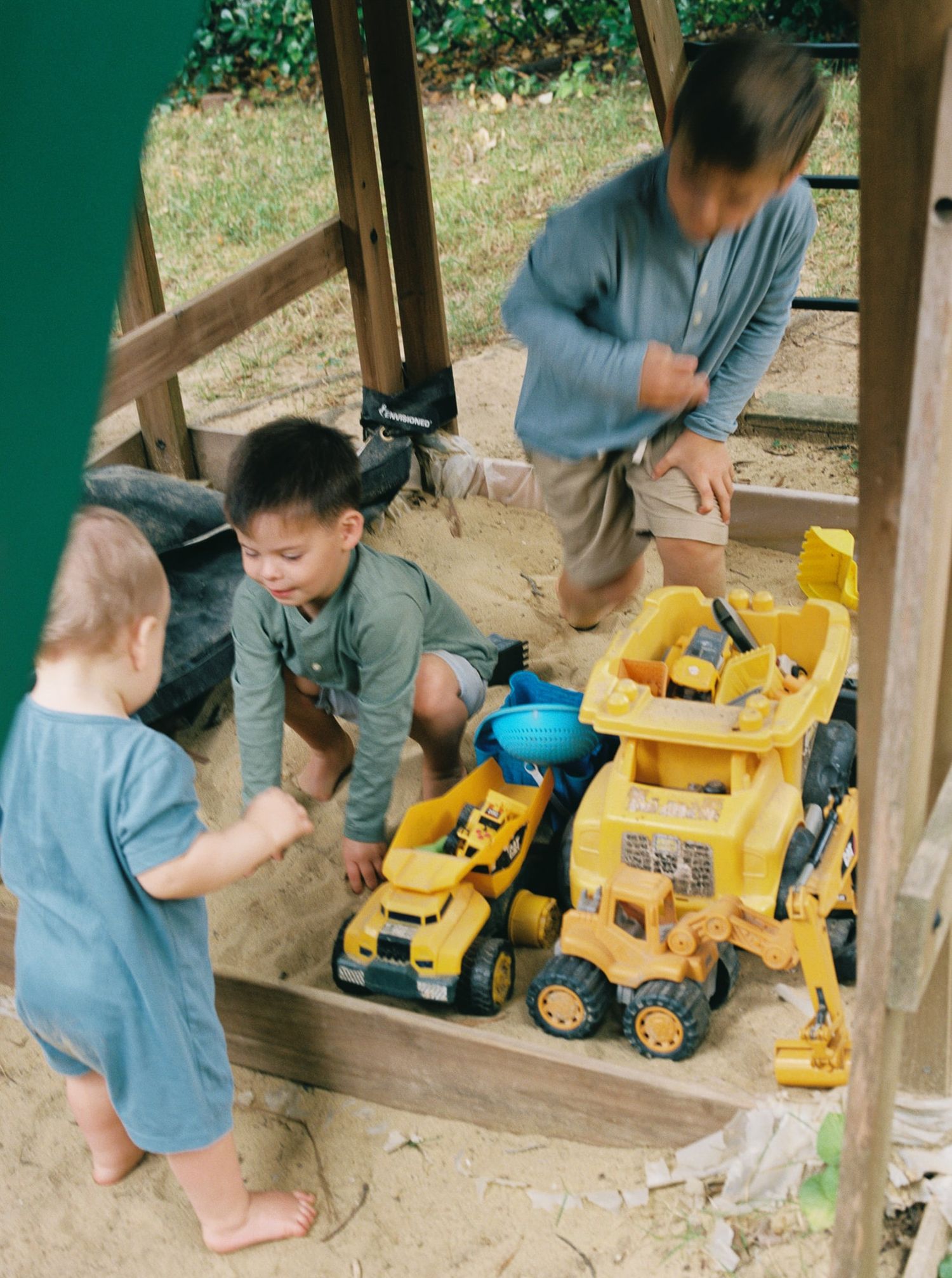 Three young children play in a backyard sandbox with yellow toy construction trucks, including a bulldozer and dump truck, on a sunny day.