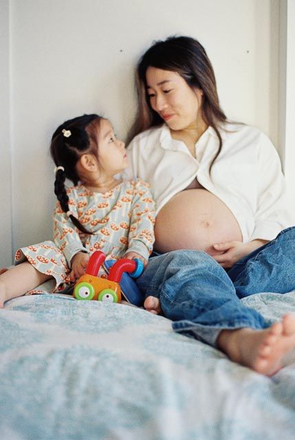 Pregnant Asian woman sitting on bed with young daughter holding colorful toy, sharing tender moment together.