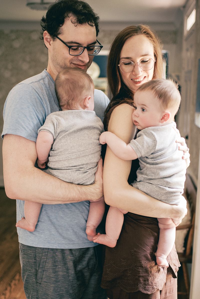 A young couple wearing glasses holds their two babies indoors, smiling warmly in a cozy home setting.