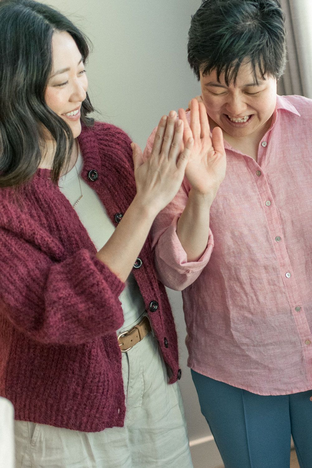 Two Asian women smiling and holding hands together indoors, wearing cozy knit sweaters in warm tones.