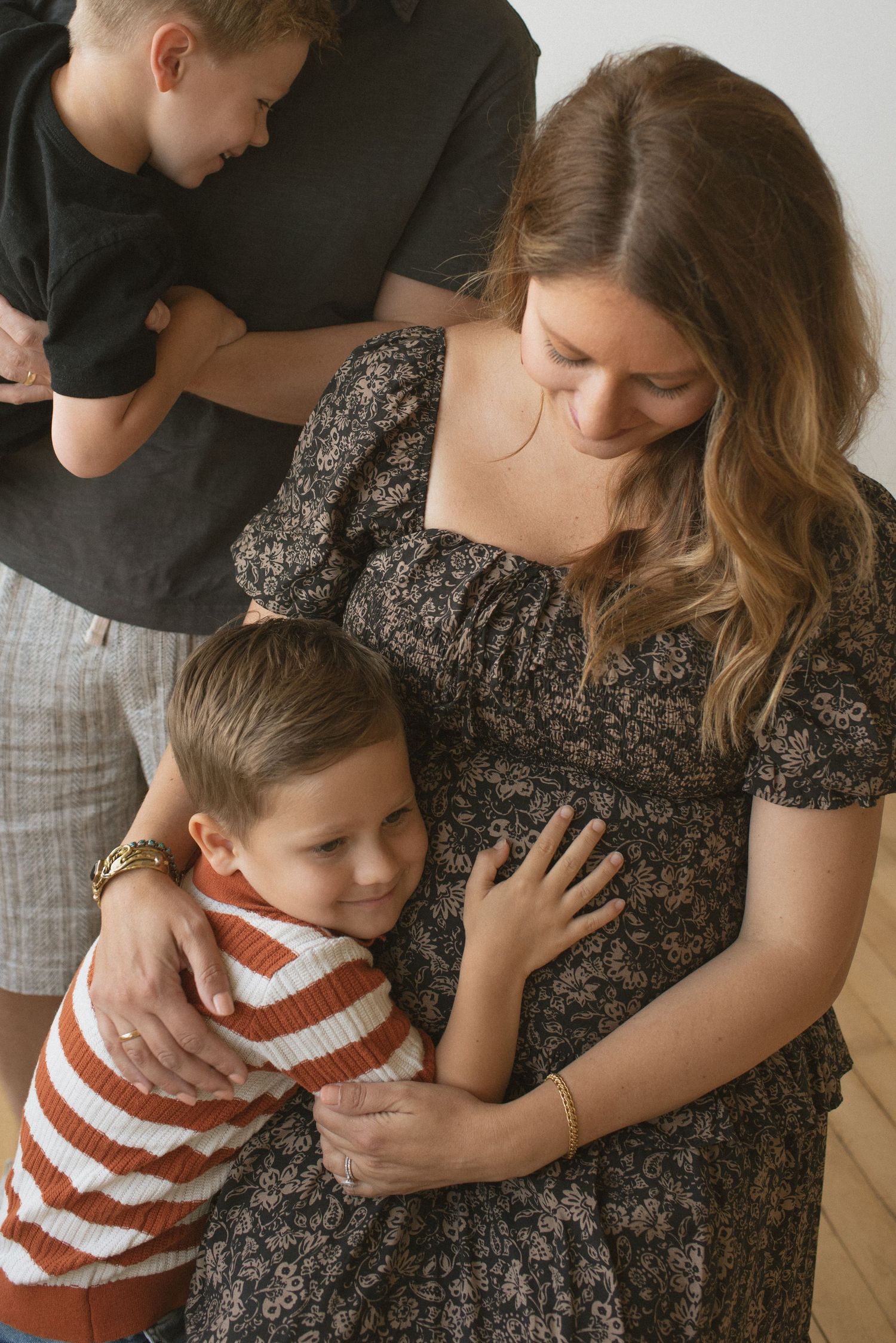 Sweet family moment as two children embrace pregnant belly in floral dress during intimate indoor maternity photoshoot.