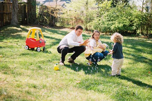 Family playing with bubbles in a sunny backyard with a Little Tikes toy car on green grass.