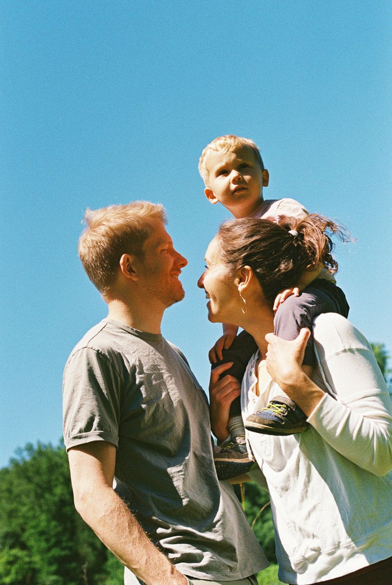 Happy family with toddler on mother's shoulders smiling under a clear blue sky.