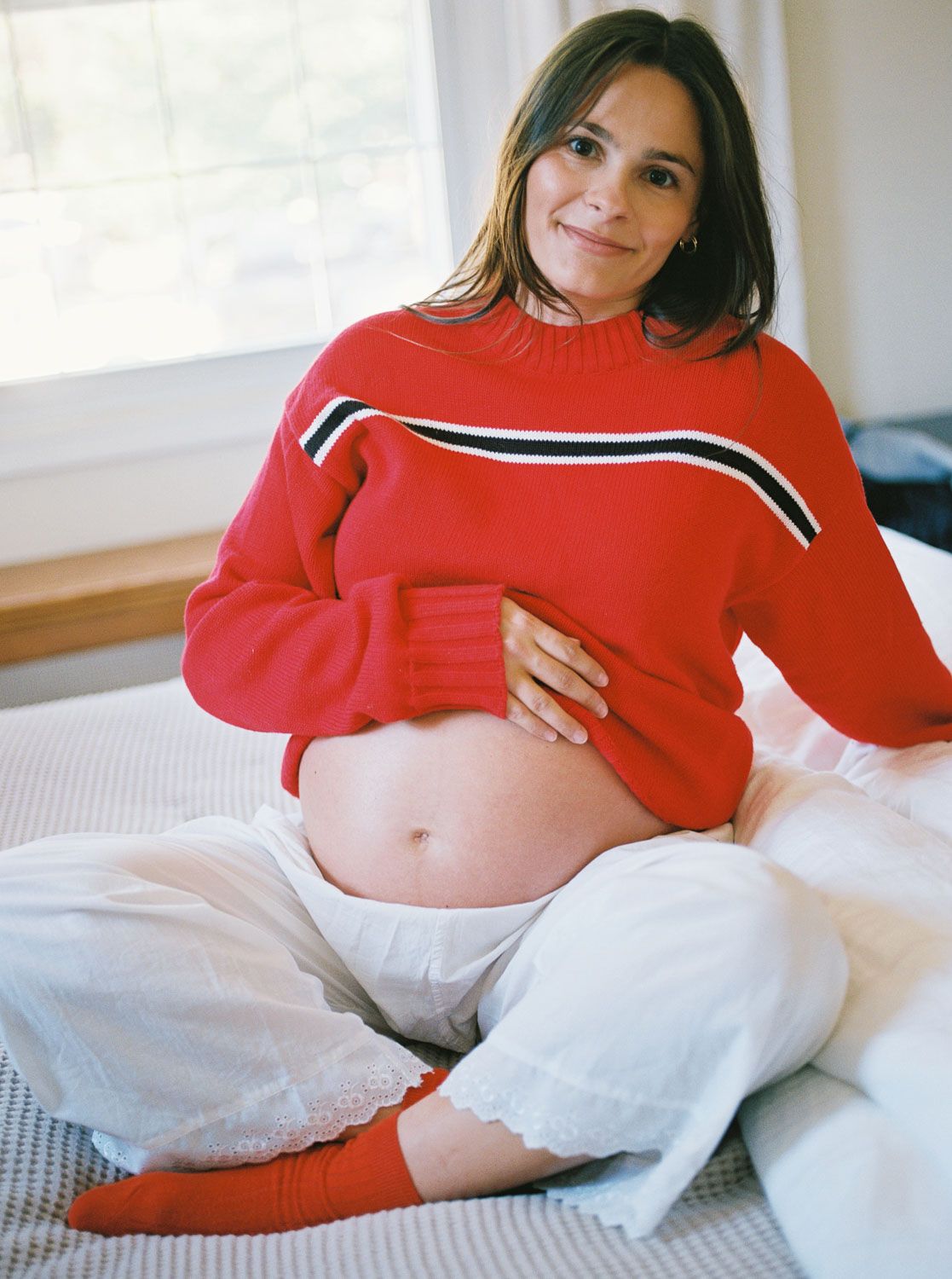 Smiling pregnant woman in a red sweater and white pants sits on a bed, gently cradling her bare belly beside a sunlit window.