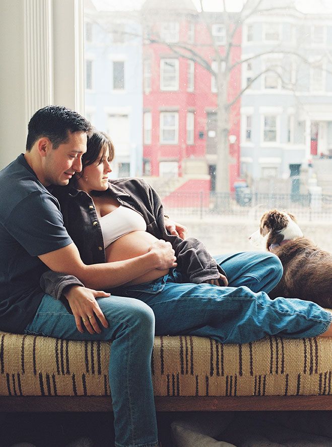 A couple relaxes together on a couch by a window with a view of red brick buildings outside.