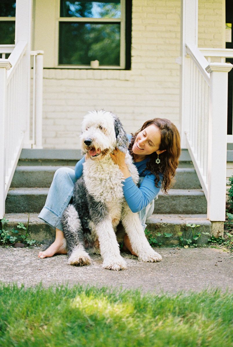 Woman sitting on porch steps hugging large fluffy Old English Sheepdog outside white brick home.