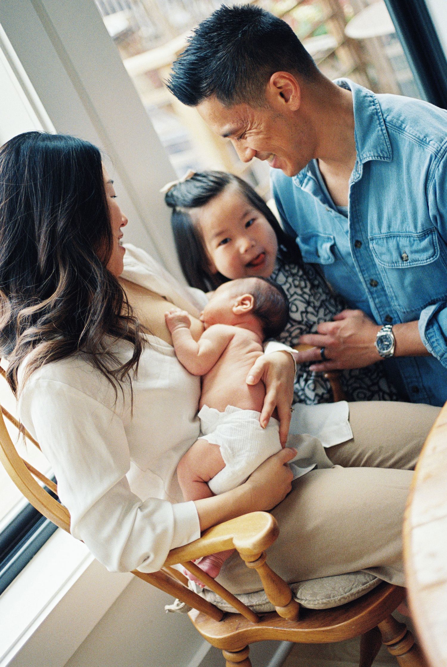 Asian family admiring their newborn baby, parents and toddler sharing a tender moment by a sunlit window.