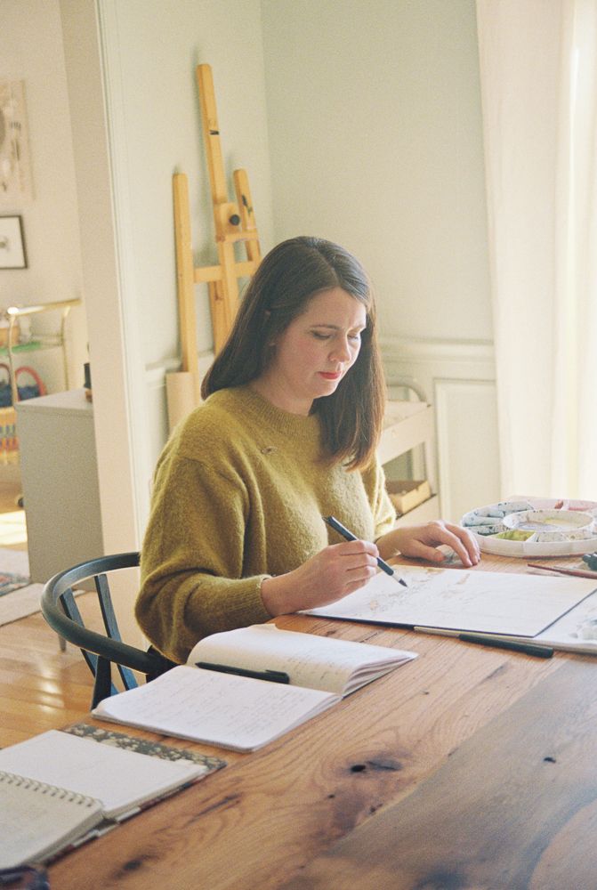 Someone works at a wooden desk with notebooks and papers spread out while wearing a mustard yellow sweater.