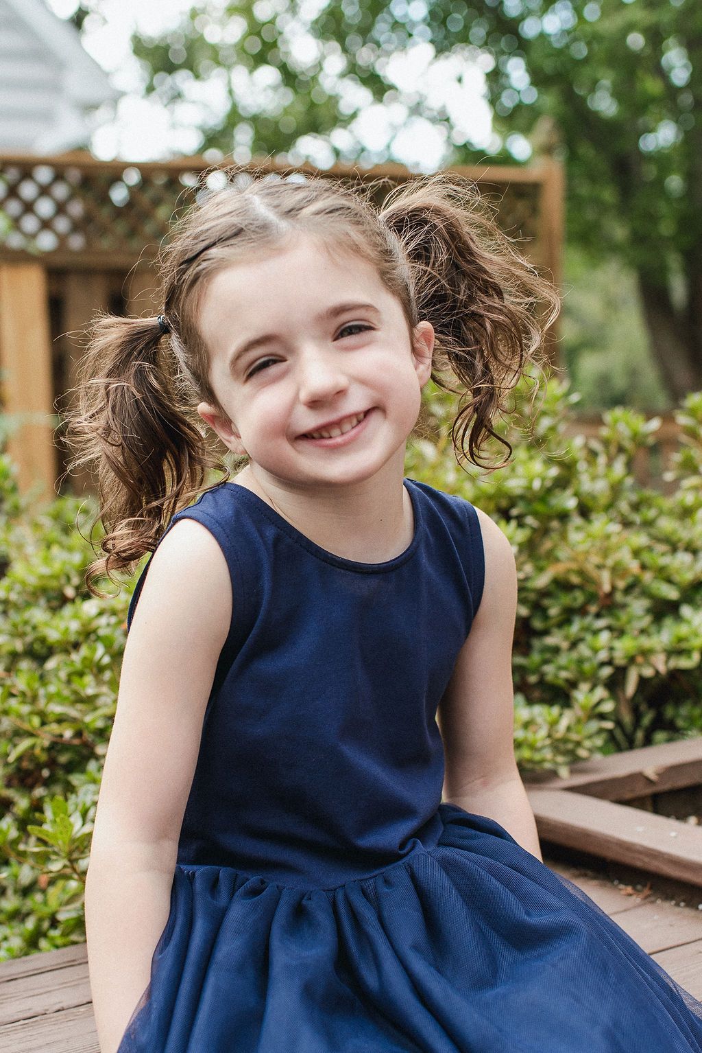Child in navy blue sleeveless dress with pigtails smiles while sitting on wooden deck in garden setting.