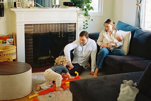 Family relaxing in cozy living room with fireplace, parents watching toddler play with colorful toy on floor.