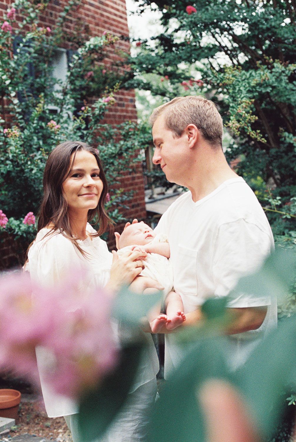 A couple in white clothing tenderly holds their newborn baby outdoors among lush greenery and pink flowers.
