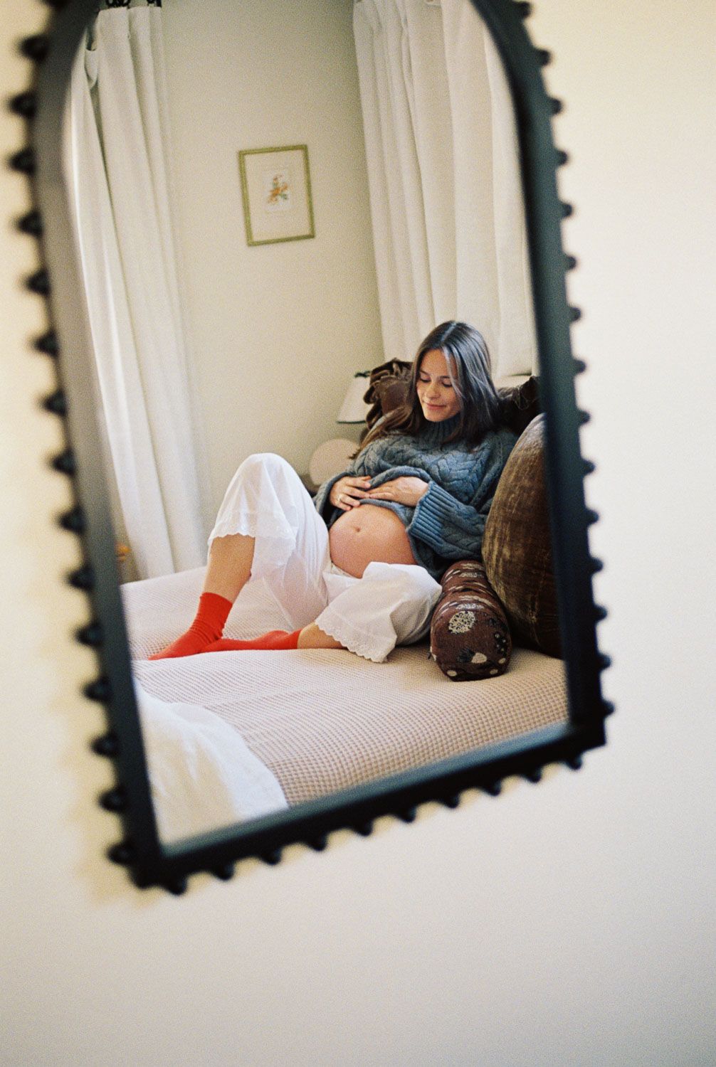 Pregnant woman in white pants and red socks sitting on a bed, reflected in a decorative mirror.