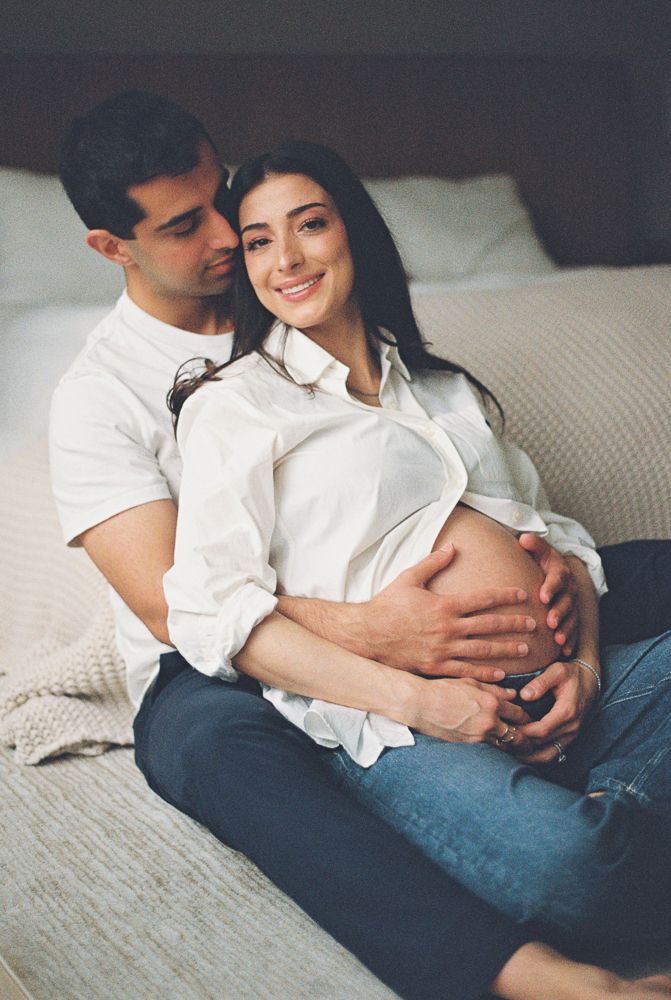 Happy pregnant couple sitting on bedroom floor, woman in white shirt showing baby bump, man embracing her from behind.