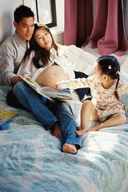 Pregnant Asian woman and man reading a book with young daughter on bed at home.
