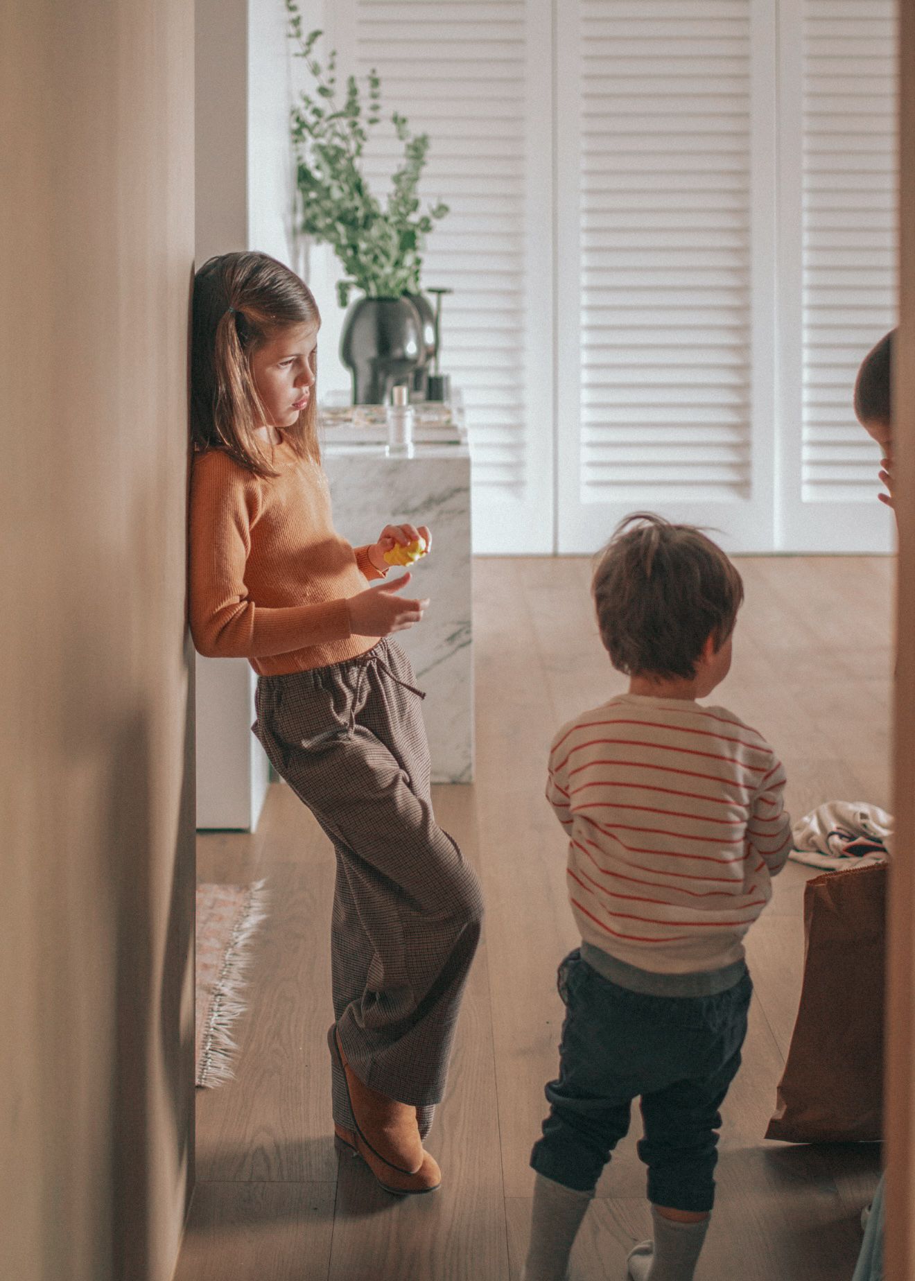 Two people stand in a sunny room with white shuttered windows as they enjoy snacks in casual attire.