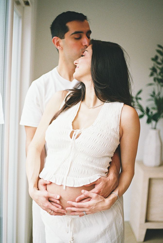A romantic couple in white clothing embraces tenderly in a bright room with plants in the background.