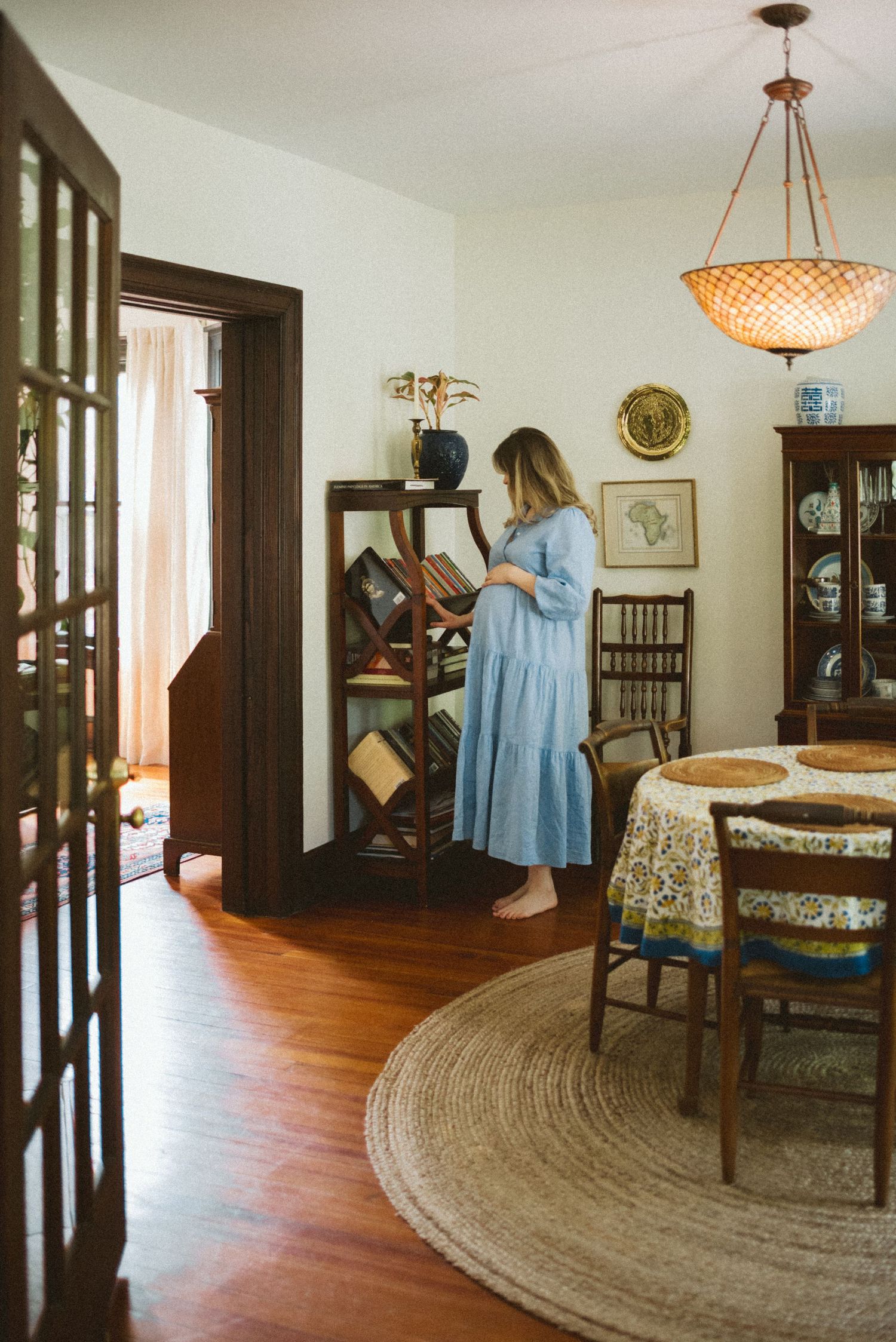 A dining room with hardwood floors features a circular jute rug, vintage furniture, and someone browsing a bookshelf.