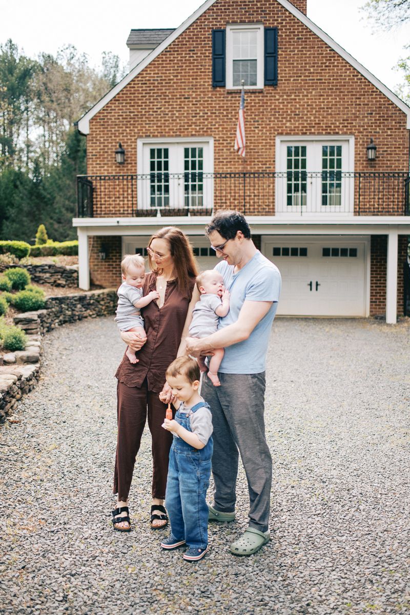 Happy family of five posing in front of a brick colonial home, parents holding twin babies with toddler standing between them.