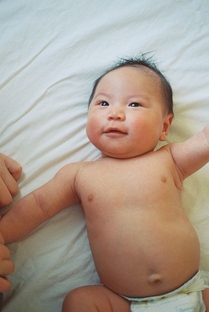 Smiling Asian baby lying on white sheets, wearing a diaper, with arms outstretched and dark hair.