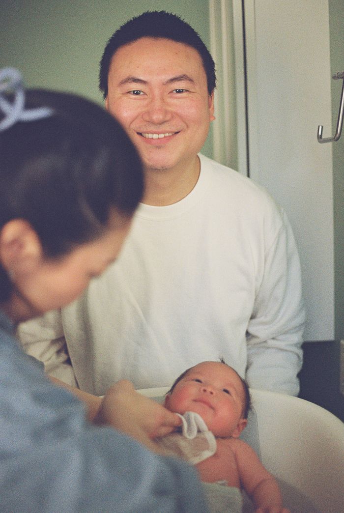 Smiling Asian man in white sweater watches a newborn baby being bathed by a woman in a bright room.