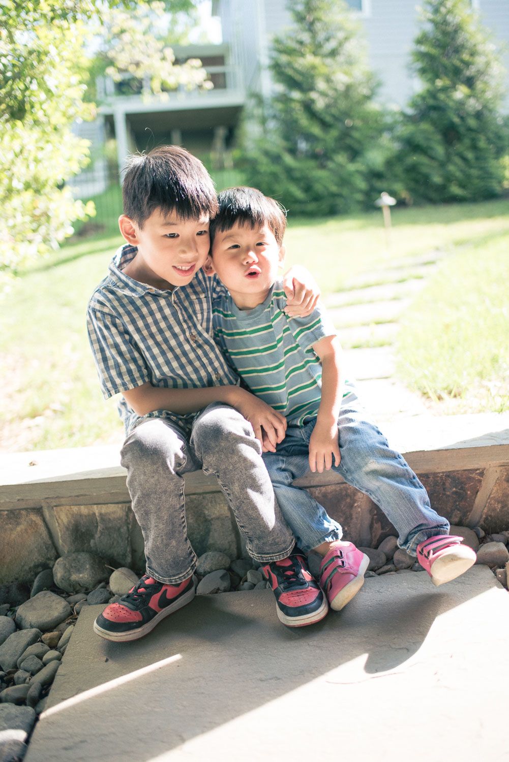 Two young Asian boys sitting together outdoors on a sunny day, smiling and hugging near a garden path.